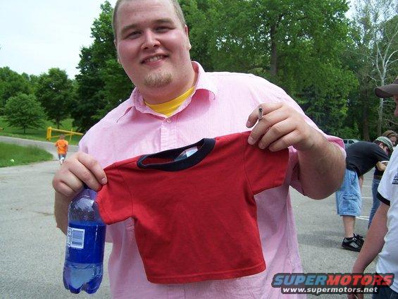 Man in pink shirt holds red t-shirt and blue water bottle; other people and cars in background