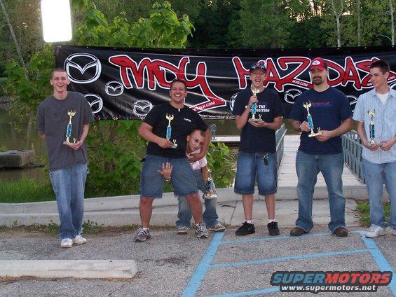 Group of five men holding trophies, standing in front of a black banner with Mazda logos and text