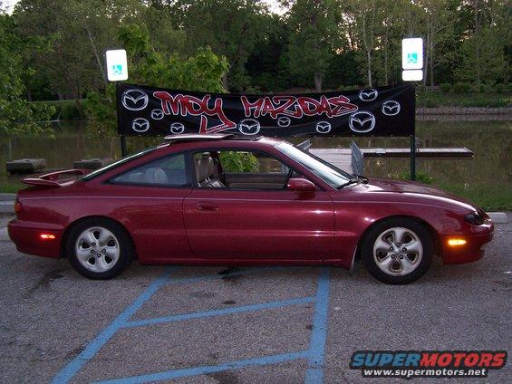 Side view of a red Mazda MX-6 coupe parked in a lot with a 'My Mazdas' banner in the background