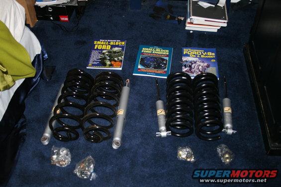 Four coil springs and four shock absorbers laid out on a dark blue carpet next to automotive repair books