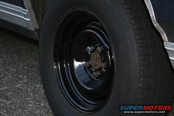 Black steel wheel with five lug nuts on a car tire. Chrome trim visible on the car body