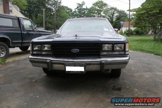 Front view of a dark blue Ford Crown Victoria with a black grille and a license plate. A dark pickup truck is parked to the left