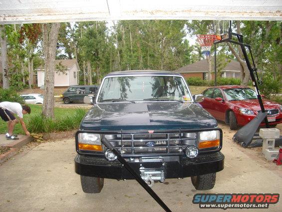 Front view of a dark green 1992 Ford Bronco with a winch and brush guard. A red sedan is parked behind it
