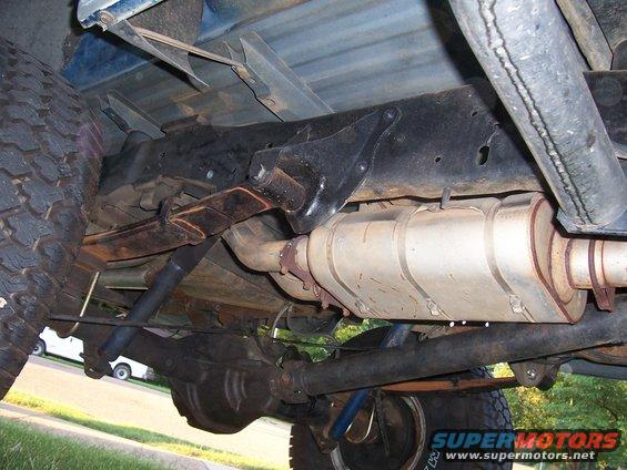 Undercarriage of a blue pickup truck, showing leaf springs, exhaust system, and rear axle. The truck appears to be from the 1980s