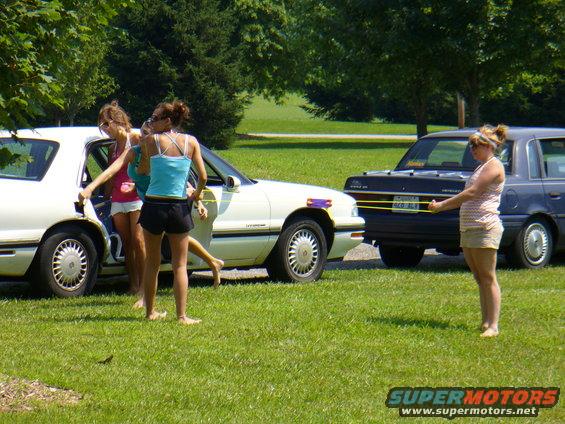Three young women by a white sedan, with a dark sedan behind it