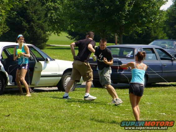People playing with water guns in a grassy area with two cars parked nearby