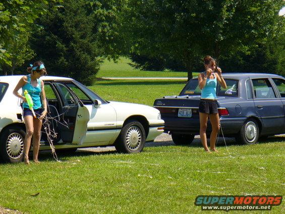 White sedan with open door and blue sedan parked on grass, two women present