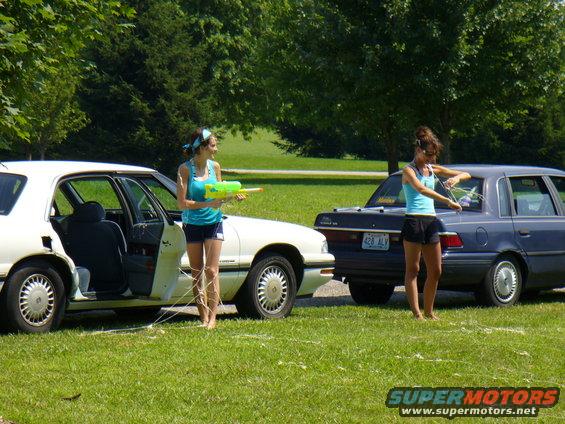 Two women stand by cars in a grassy area, one holding a water gun