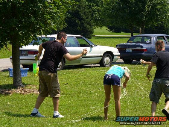 People having a water gun fight in a park with two cars in the background