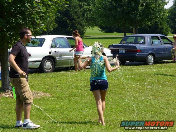 People at an outdoor gathering with two cars in the background. One person has something green on their head