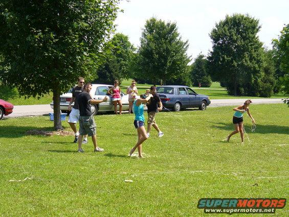 People playing in a grassy park with several cars parked in the background