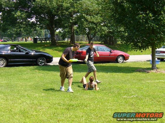Two guys with water guns aiming at a person on the grass, with a black car and a red car parked nearby