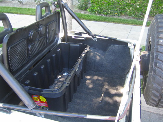 Open black storage container in the cargo area of a white off-road vehicle with a spare tire visible on the right