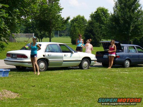 Two sedans, a white one and a blue one, parked on grass. People are standing around them