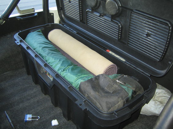 Open black storage box in vehicle interior containing green sleeping bag and rolled tan mat