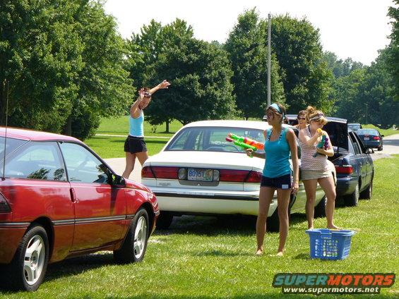 Red coupe parked on grass, side view. White sedan behind it