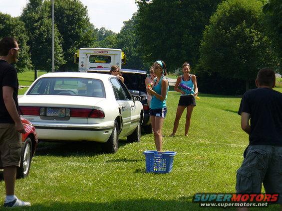 White sedan parked on grass, people with water guns around it