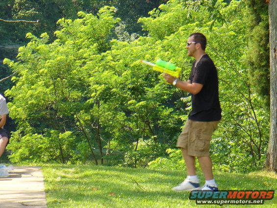 Man in black t-shirt and khaki shorts holding a yellow and green water gun, standing on grass