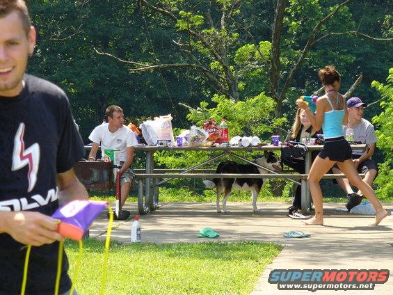 People gathered at a picnic table with a dog, a man in the foreground holds a toy