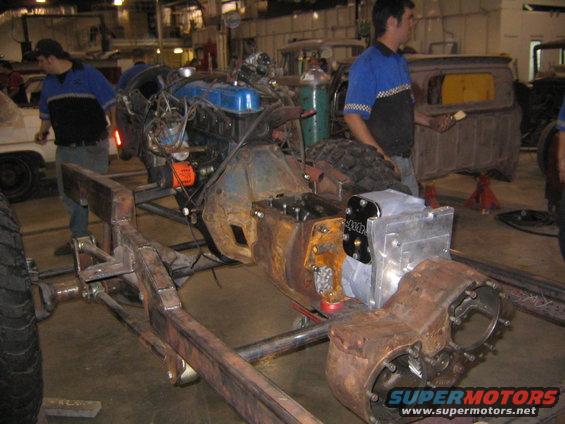 Rusty vehicle frame with blue engine, transmission, and differential assembly. Men in blue shirts work nearby
