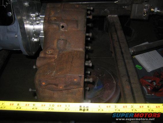 close-up of a rusty metal component bolted to a silver part, with a yellow measuring tape in front