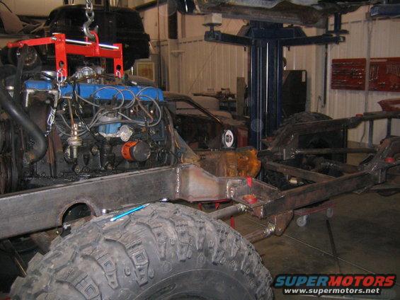 Engine being lowered onto a vehicle frame in a workshop. A large tire is in the foreground