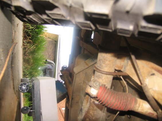 Underside view of vehicle suspension, showing a shock absorber with a red boot and rusty metal components