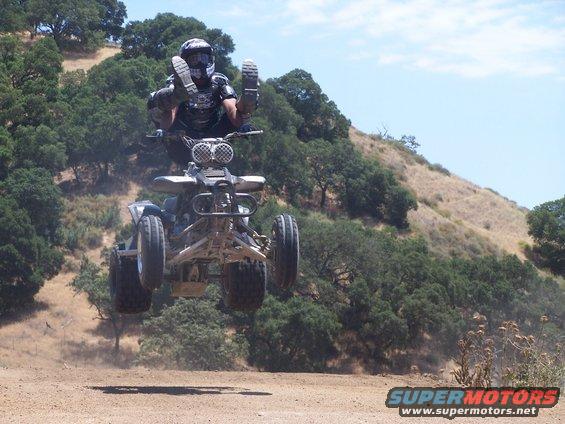 Quad bike airborne with rider, front view. Dusty terrain and trees in background