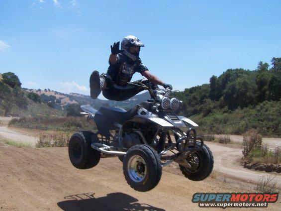 Person in mid-air on a four-wheeler ATV, waving. Dirt track and hills in background