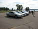 2003 Ford Crown Victoria - Several sedans, including a silver 2003 Ford Crown Victoria, are parked in a lot near a body of water.