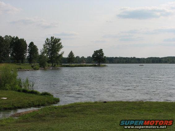 Lake with grassy shore, trees, and a small boat on the water under a cloudy sky