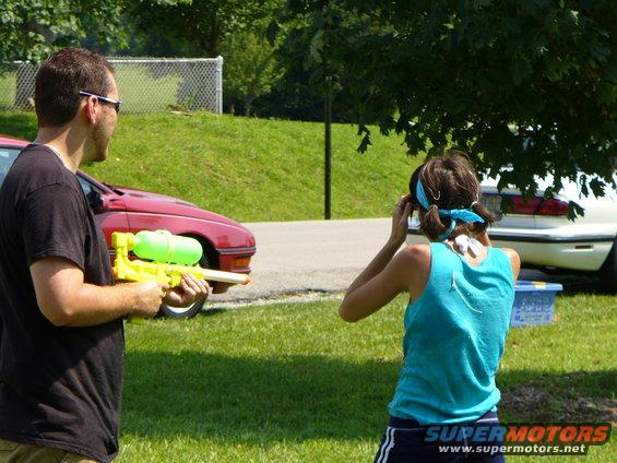 Man holding yellow water gun, woman in blue tank top, red car, white car in background