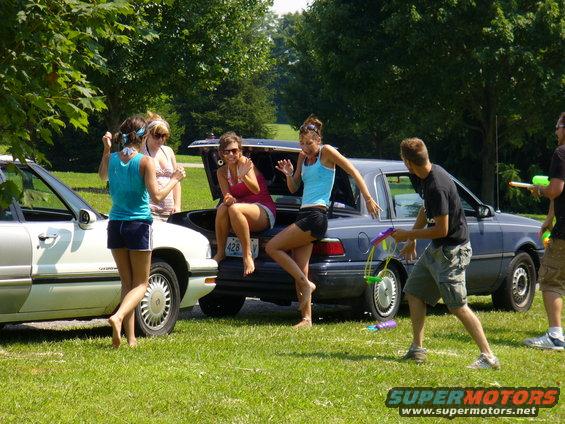 People hanging out by cars, some sitting in a trunk, one holding a water gun