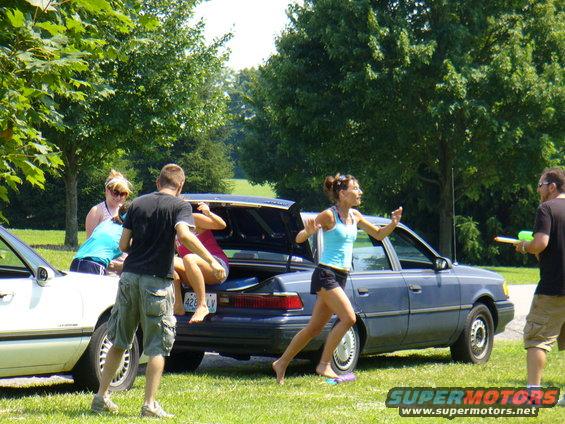 Blue sedan with open trunk, people interacting, one person in trunk, another running. License plate 426 LV
