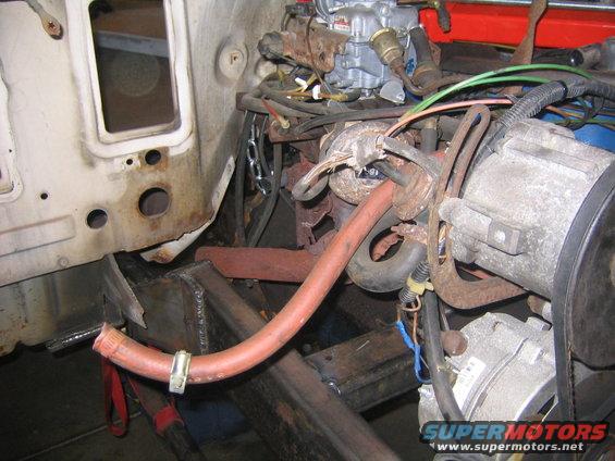 Engine bay with rusty exhaust manifold, carburetor, and alternator. A red hose is clamped to a metal bracket