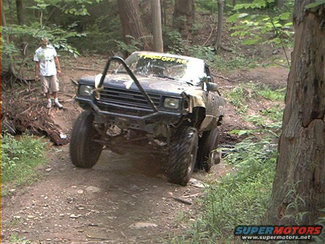 Off-road truck with a brush guard navigating a wooded trail. A person stands on the side of the path