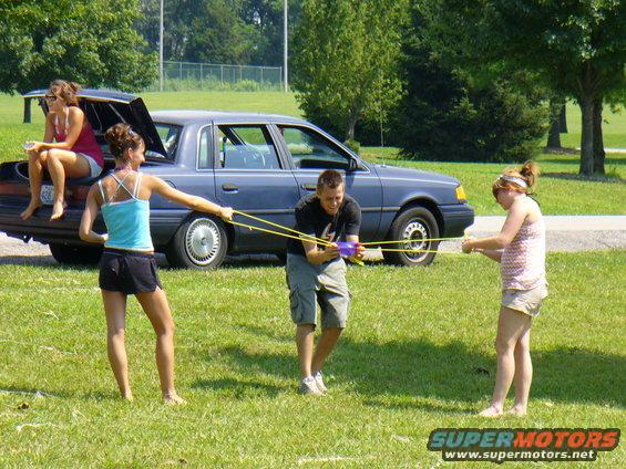 Four people in a grassy area, one sitting in the trunk of a blue sedan, three others pulling on a yellow rope. The sedan appears to be from the early 1990s