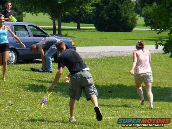 People playing with water guns in a grassy area with a blue car in the background