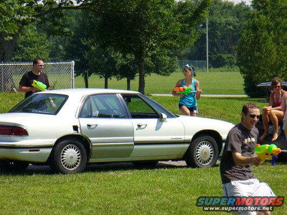 White sedan side view, people with water guns in a park