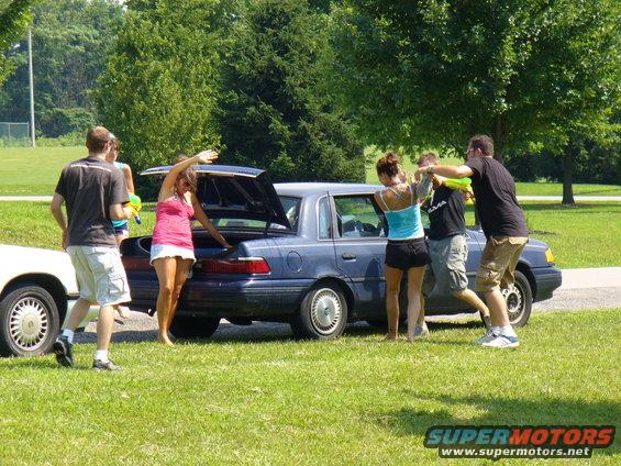 Blue sedan with trunk open, people gathered around, one white car visible on left