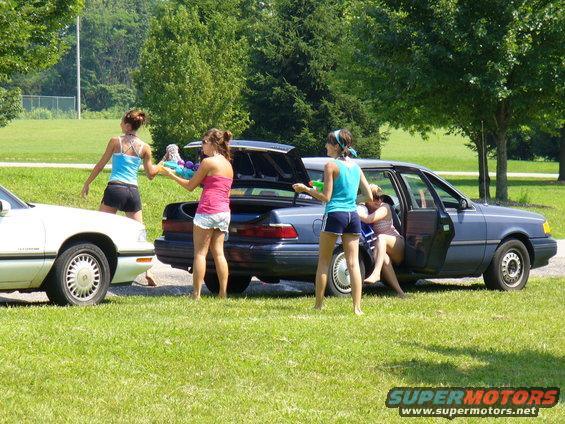 Two women with water guns by a dark sedan with an open trunk; another car is parked to the left
