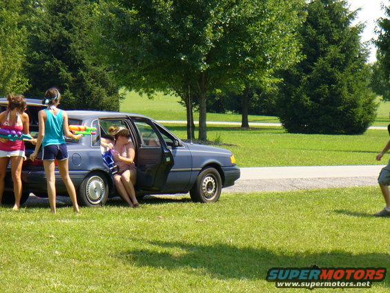 Blue sedan with open doors, people with water guns in and around it, on a grassy area