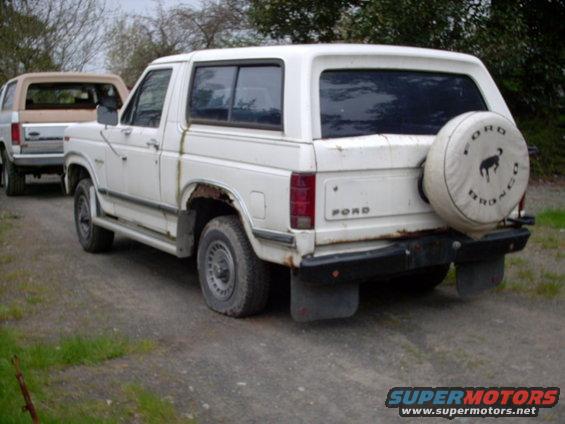White 1986 Ford Bronco parked on a dirt road, another Bronco behind it. Spare tire cover has Bronco logo