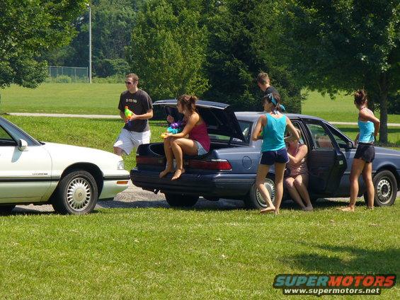 Two cars parked on grass with people standing and sitting around them