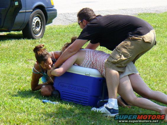 People wrestling on a cooler next to a blue car