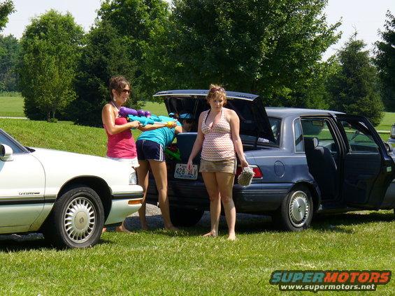 Two women with water guns stand by a blue sedan with an open trunk, next to a white car