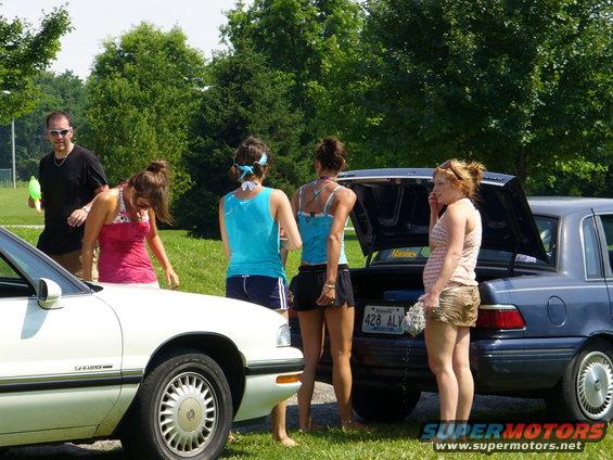 Group of people by a white car and a blue sedan with open trunk. License plate reads 428 ALV
