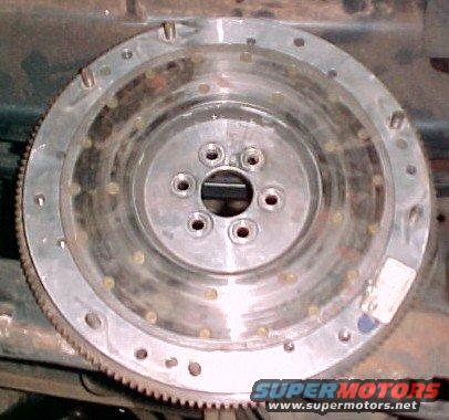 Metal flywheel with bolt holes and gear teeth around the edge. Supermotors.net logo at bottom right
