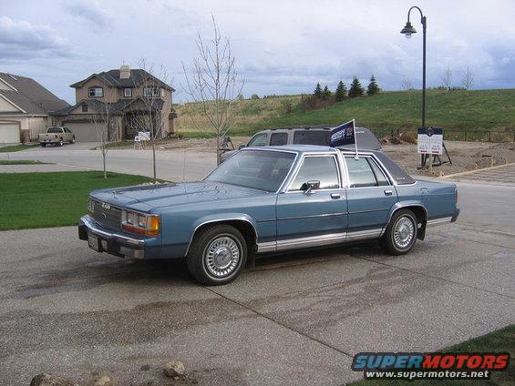 Blue sedan parked on a driveway, with a house and trees in the background. A flag is attached to the roof