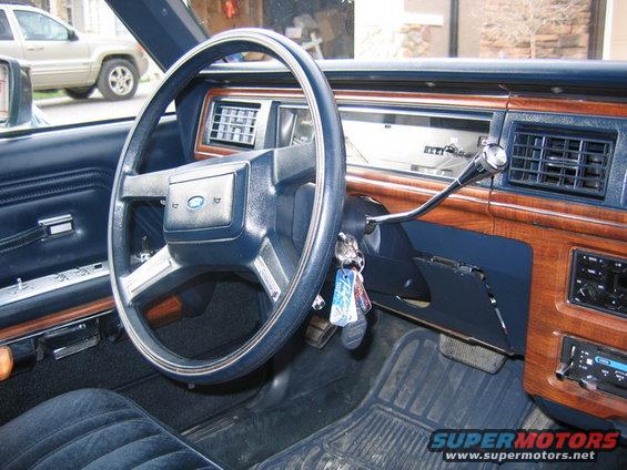 Car interior with blue velour seats, wood trim dashboard, and Ford steering wheel. A beige SUV is visible outside